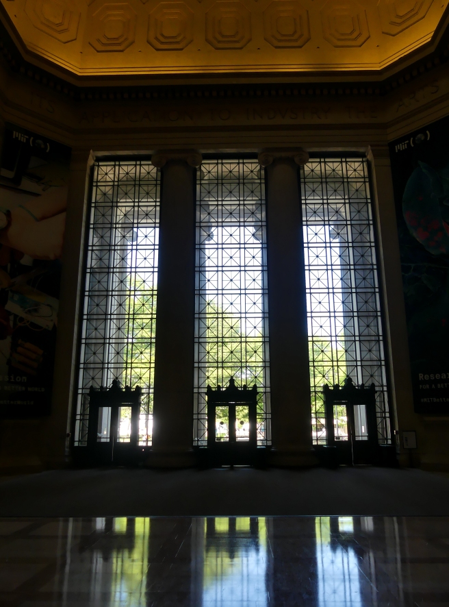View from inside a dark hall with tall ceilings, looking out through three tall windows onto a sunny day.