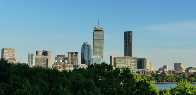 Boston skyline with trees in the foreground and blue sky above it.